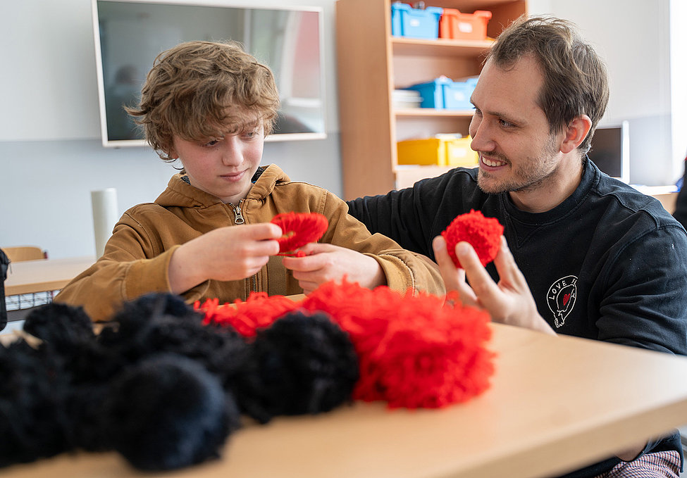 Junge fertigt roten Pompon, Betreuer lächelt und hält einen weiteren; Tisch mit schwarzen und roten Bommeln, Klassenraum im Hintergrund.
